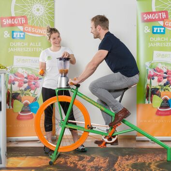 Mannsitzt auf Smoothiebike, Frau sichert den Deckel auf dem Mixer
