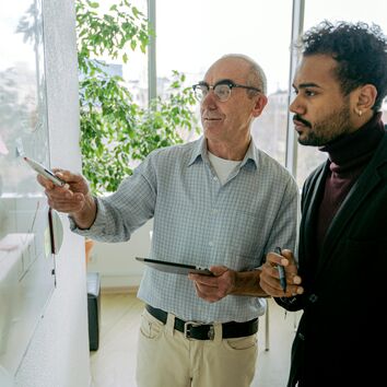 Junger Mann wird von älterem Mann vor einer Tafel stehend angeleitet